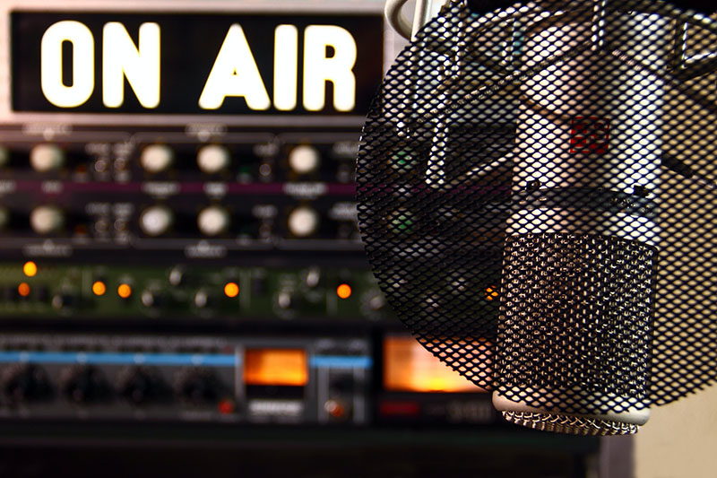 A close-up picture of a microphone in a recording studio with the words “on air” in the background. 