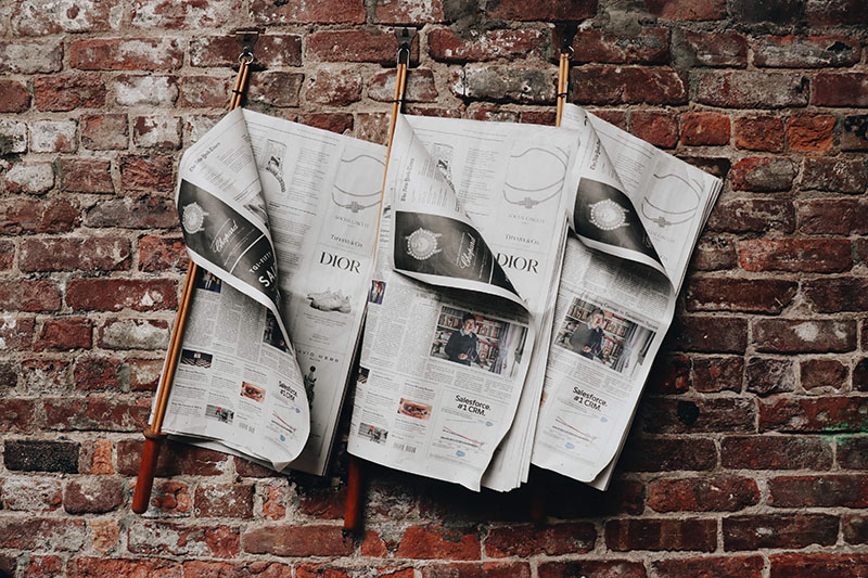 Three newspapers hanging on clips on a brick wall.