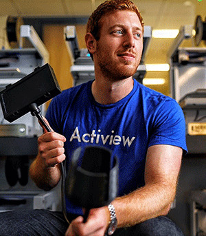 A picture of accessibility app Actiview founder Alex Koren, wearing a blue Actiview t-shirt and holding some of the company's accessibility equipment.