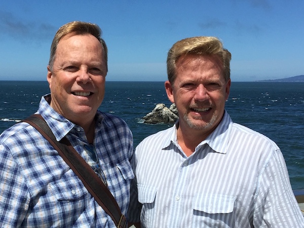 Photo of Terry Wicks and his husband Michael Draper, standing side by side, smiling, with the ocean in the background.