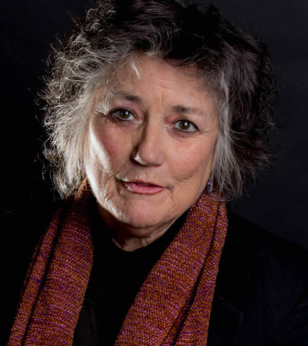 Author Terry Galloway, a woman with short, messy, and stylish gray hair smiles at the camera, wearing a black shirt with a red scarf.