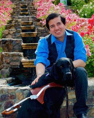 Belo Miguel Cipriani sits in front of a garden waterfall surrounded by flowers. He's wearing a blue button up shirt, a black vest, and black pants. His guide dog, Oslo, a black lab, sits at his feet wearing a harness.