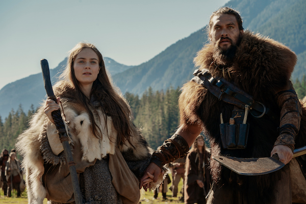 Actors Jason Momoa and Hera Hilmar from the show See stand together, draped in furs in a rustic, mountain setting.