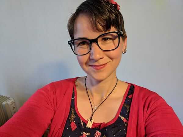 Filmmaker Karina Sturm smiles at the camera, wearing a red cardigan and black-framed glasses.