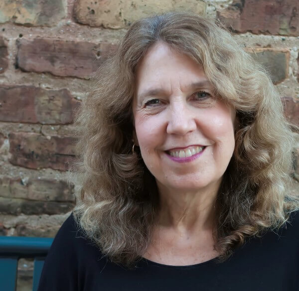 Author Lisa M. Schab smiles at the camera, standing in front of a brick wall. She has shoulder-length, curly, blonde hair, and is wearing a black shirt.