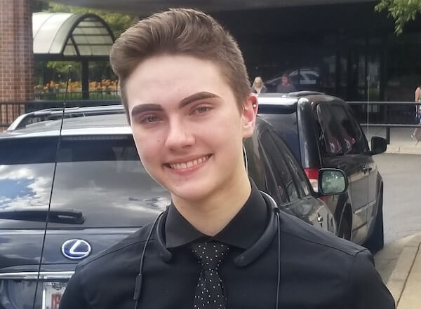15-year-old Sebastian Duesing smiles at the camera. He's wearing a black collared shirt and a black tie with specks of patterned white.