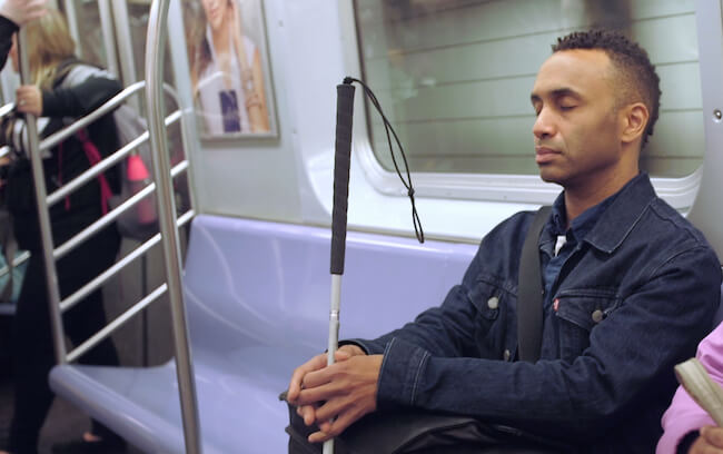 Vision Portraits director Rodney Evans sits on a subway train with his eyes closed holding a white cane.
