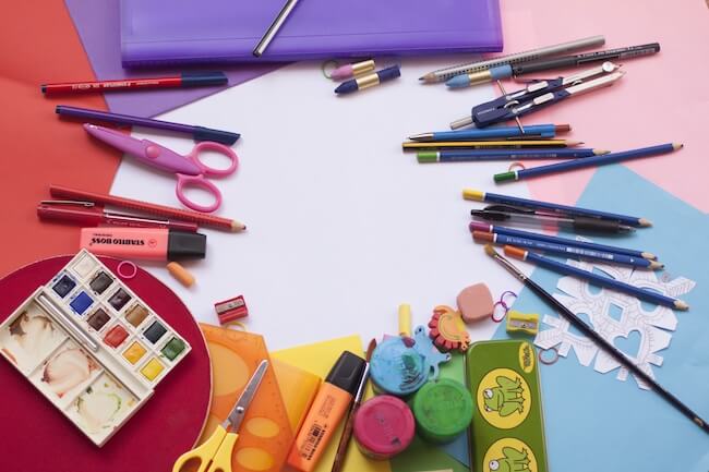 An array of school supplies spread on a table, including painting and drawing supplies, scissors, stamps and paper.