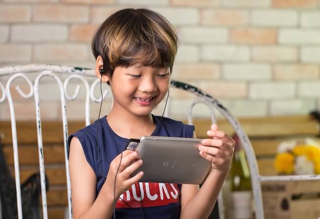 A boy wearing headphones is looking at a tablet smiling.